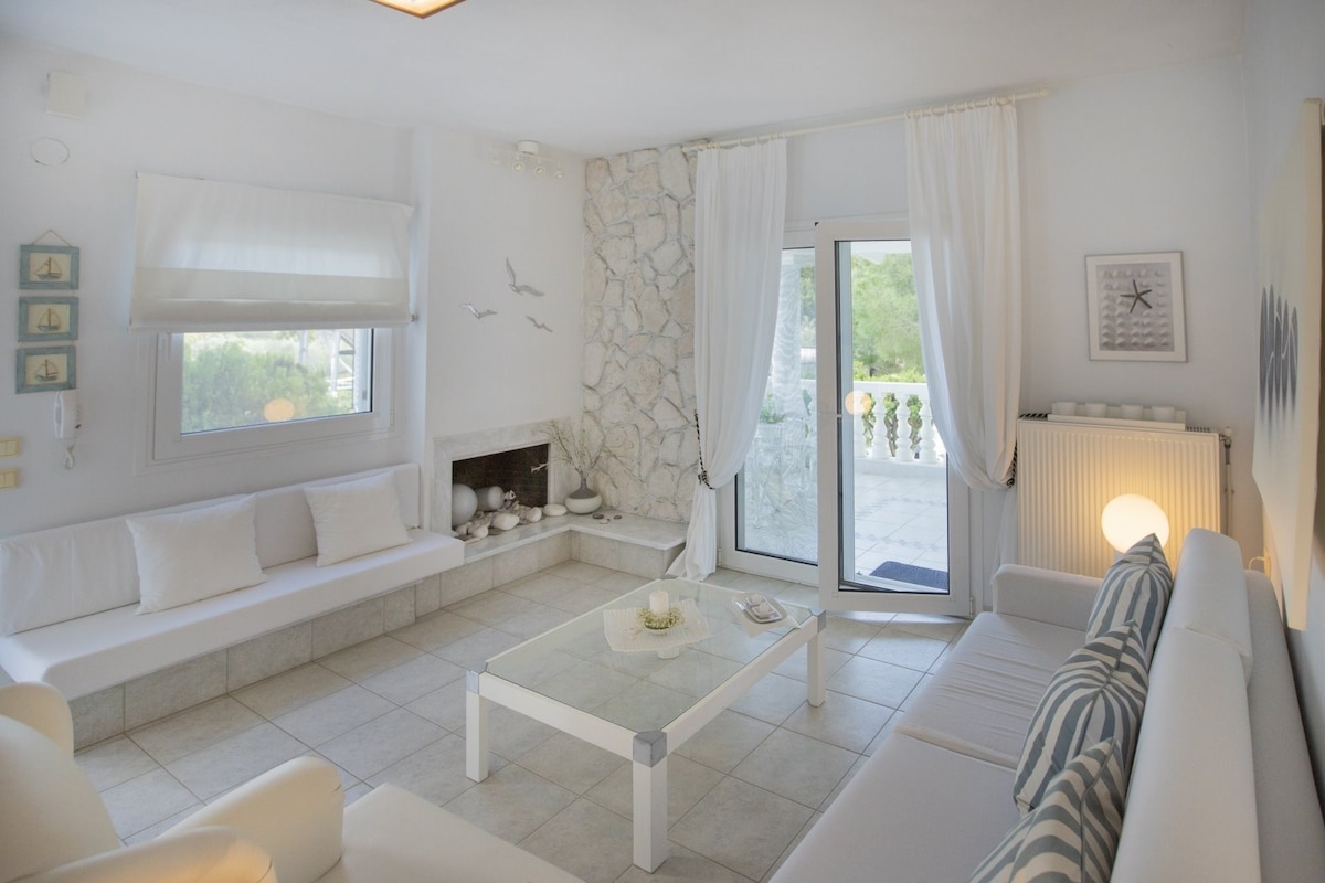 A light-filled living area features comfortable seating arranged around a low coffee table. A stone wall adds texture, while sheer white curtains soften the sunlight. Large glass doors open to the outdoors, connecting the space with the lush surroundings.