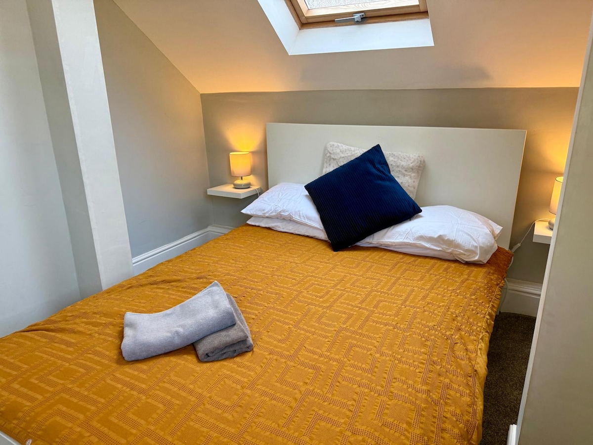 A cozy bedroom is highlighted by a warm golden bedspread and plush white pillows. A navy blue cushion adds a touch of colour against the neutral wall. Soft lighting from bedside lamps creates a relaxing ambiance, complemented by a skylight that allows natural light to filter in.