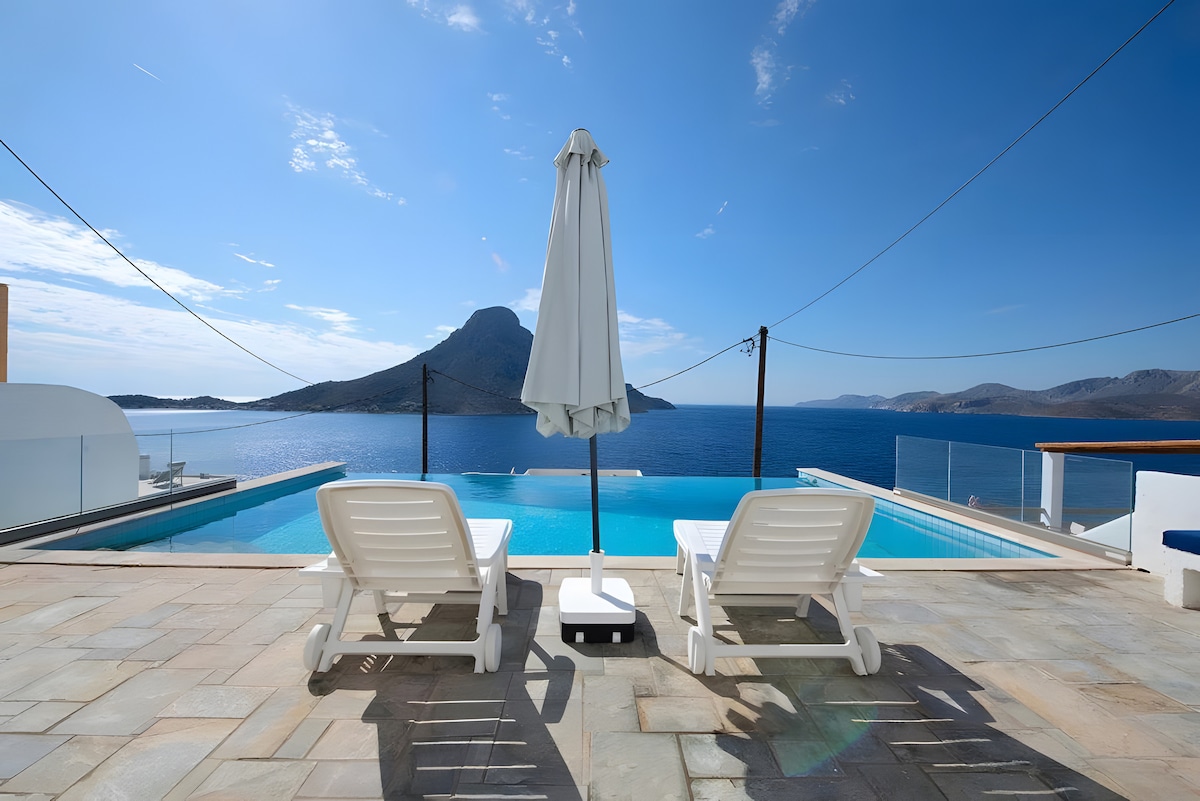 A serene outdoor area features a private pool overlooking the Aegean Sea, complemented by two white lounge chairs and a large umbrella providing shade. A scenic view of a nearby mountain adds to the tranquil setting, with clear blue skies reflecting on the water.