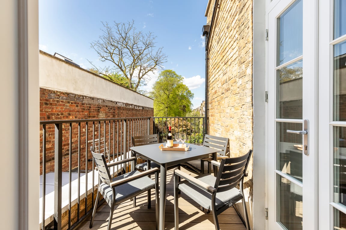 A private balcony is shown with outdoor seating for four around a sleek dining table. Light grey wooden flooring complements the greenery visible in the background. The space is bordered by a modern railing and features a clear sky above.