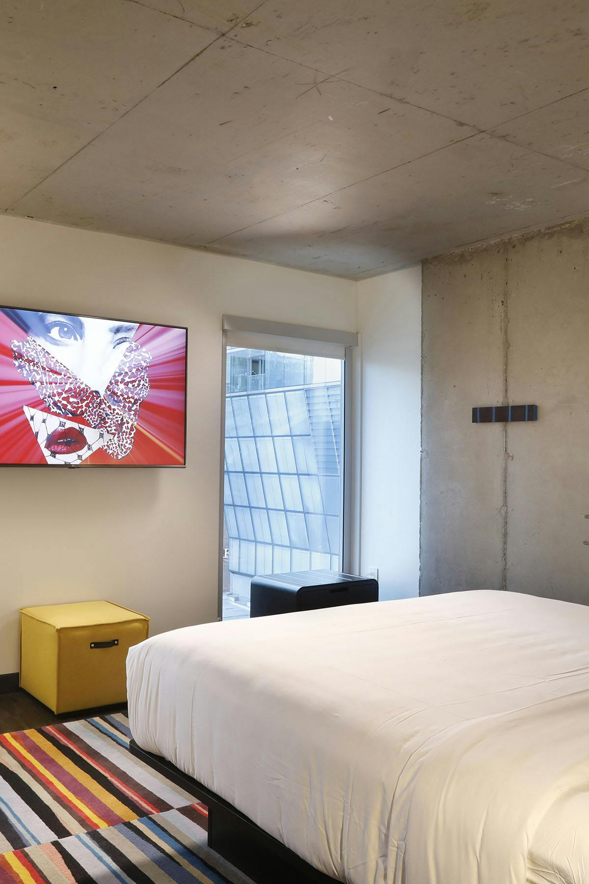 A modern hotel room features a spacious bed with crisp white linens. An oversized TV is mounted on the wall, displaying vibrant artwork. A yellow storage cube is positioned next to the bed, and a colorful striped rug adds texture to the dark floor.