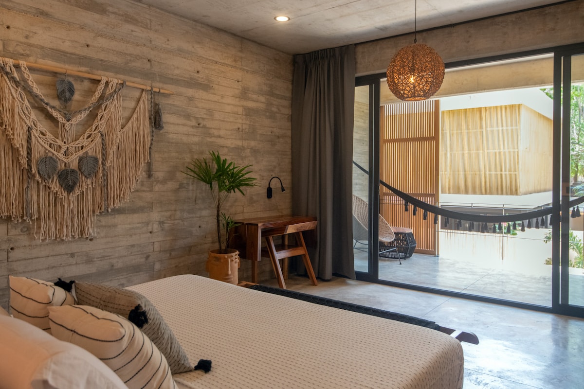 A spacious room features a comfortable bed adorned with decorative pillows. Natural light fills the space through large glass doors, leading to a private terrace. A macramé wall hanging adds texture, while a small wooden desk and a potted plant are positioned nearby.
