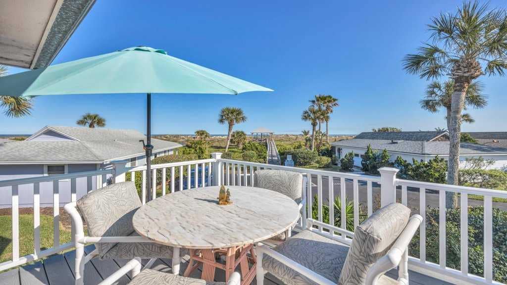 A sunlit outdoor dining area is presented, featuring a round marble table surrounded by white chairs. A large aqua umbrella offers shade, while palm trees and distant rooftops create a serene backdrop under a clear blue sky.