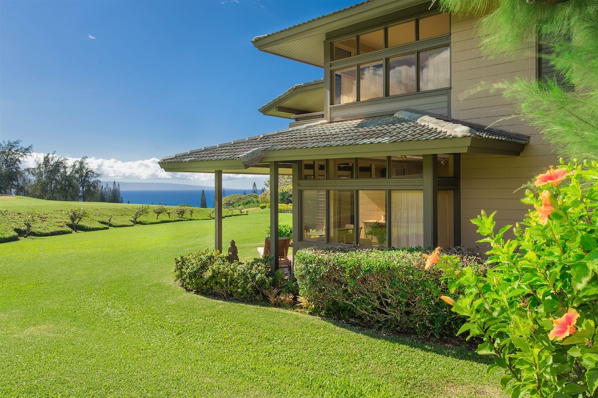 The exterior of the townhouse features a landscaped yard with well-maintained grass and vibrant tropical plants. The structure showcases large windows that invite natural light, while a covered porch area offers a shaded spot to enjoy outdoor views, including glimpses of the Pacific Ocean.