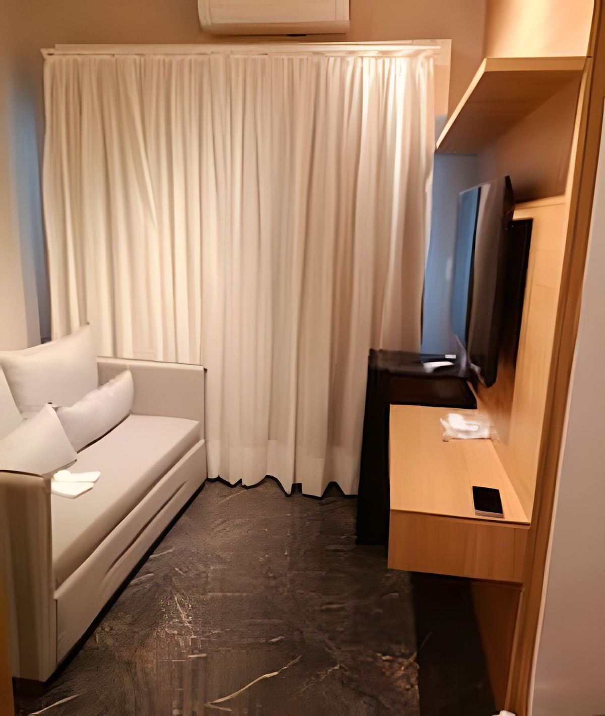 A cozy seating area is featured, equipped with a comfortable gray sofa positioned beside a large window covered by sheer white curtains. A wall-mounted television and a wooden desk add functionality to the space, while a small refrigerator is situated nearby.