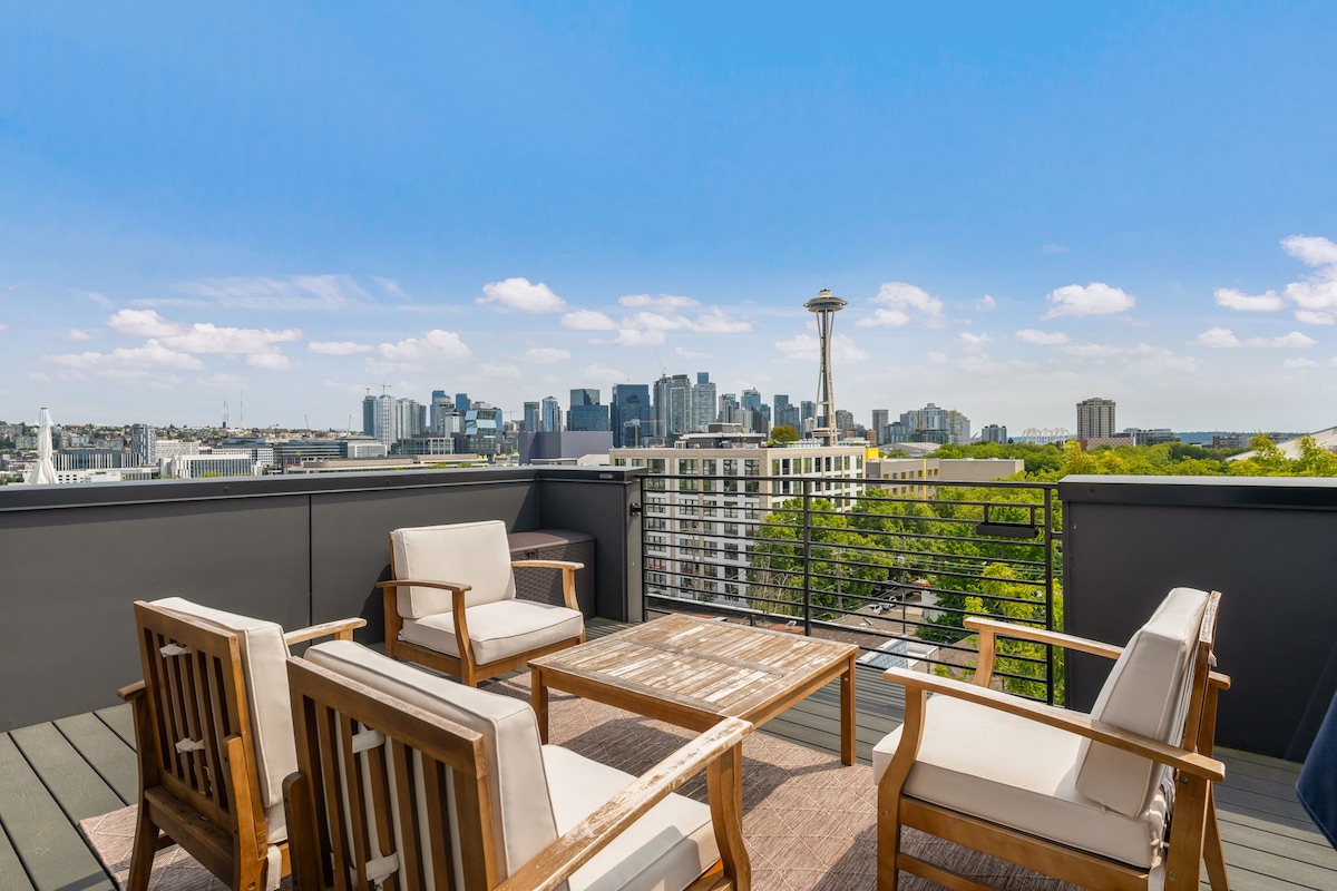 An outdoor patio features wooden furniture with cushioned seating arranged around a central table. The expansive view displays Seattle's skyline, including the Space Needle, under a clear blue sky with scattered clouds.