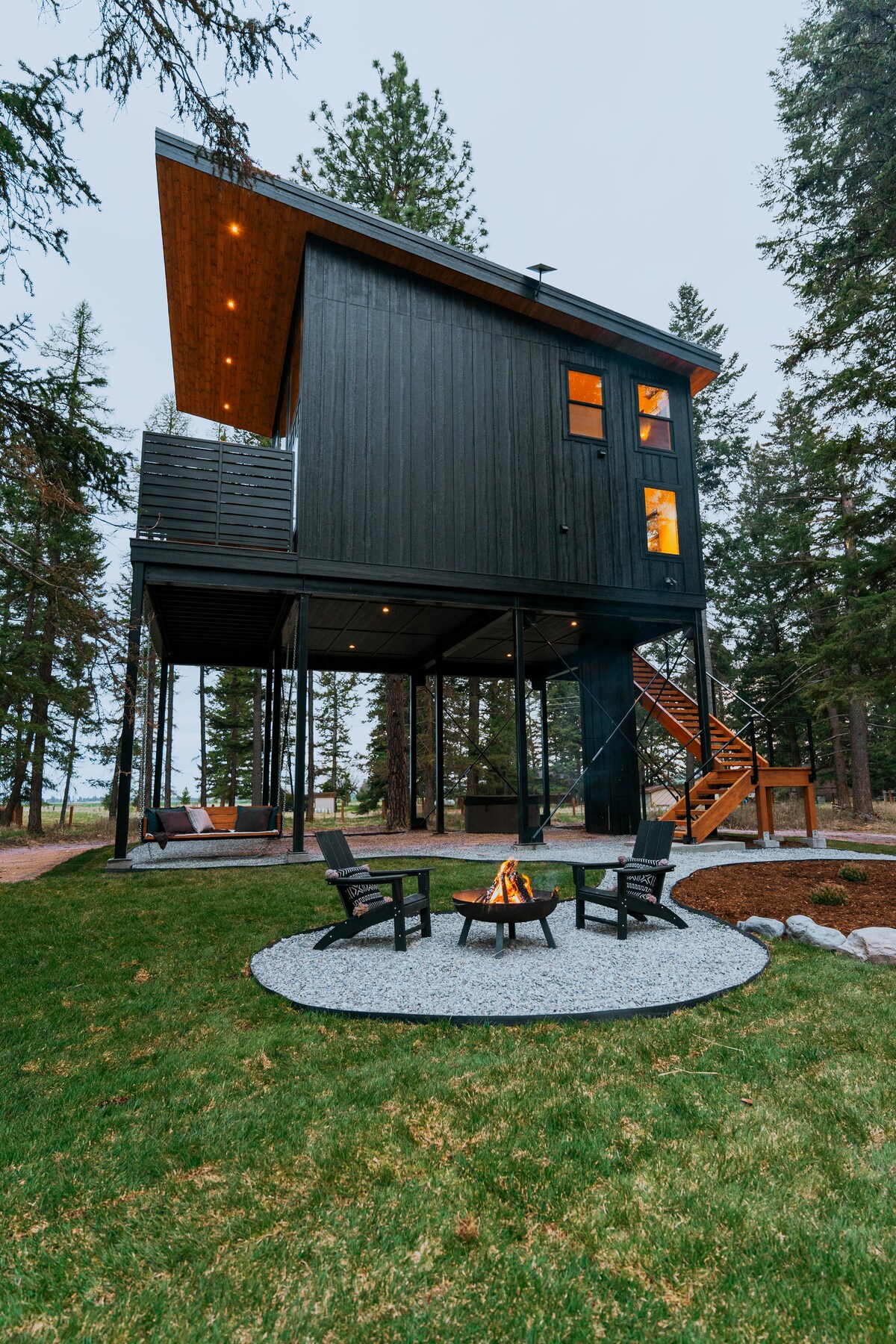 A modern treehouse stands elevated on stilts amid tall trees. Below, a fire pit surrounded by four black chairs is set on gravel, creating a cozy outdoor gathering spot. Soft lighting highlights the treehouse structure as dusk approaches.