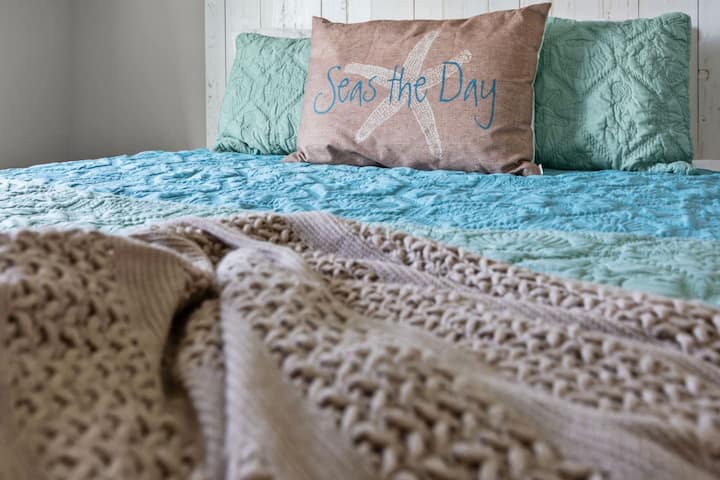 Relájate y desconecta con la comodidad de la costa. Nuestro encantador dormitorio te invita a "Seas the Day" con sus relajantes tonos verdes de mar y sus acogedores mantos de punto. Perfecto para una escapada serena.