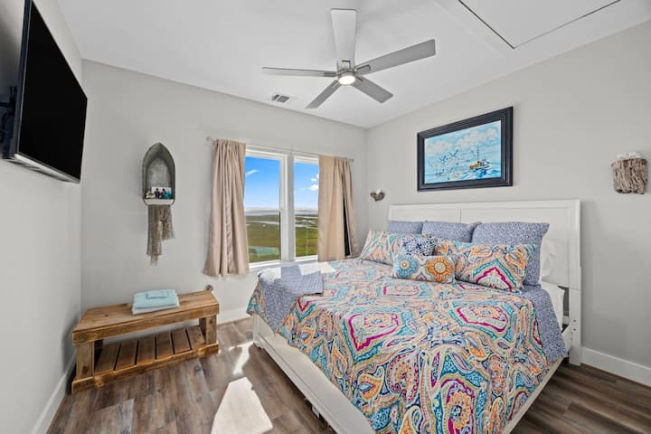 Vibrante y acogedora, esta cama tamaño king con cabecera blanca y ropa de cama colorida y intrincada aporta encanto junto al mar. La decoración náutica y un banco de madera rústico mejoran el espacio, mientras que los grandes ventanales ofrecen vistas impresionantes del paisaje abierto.