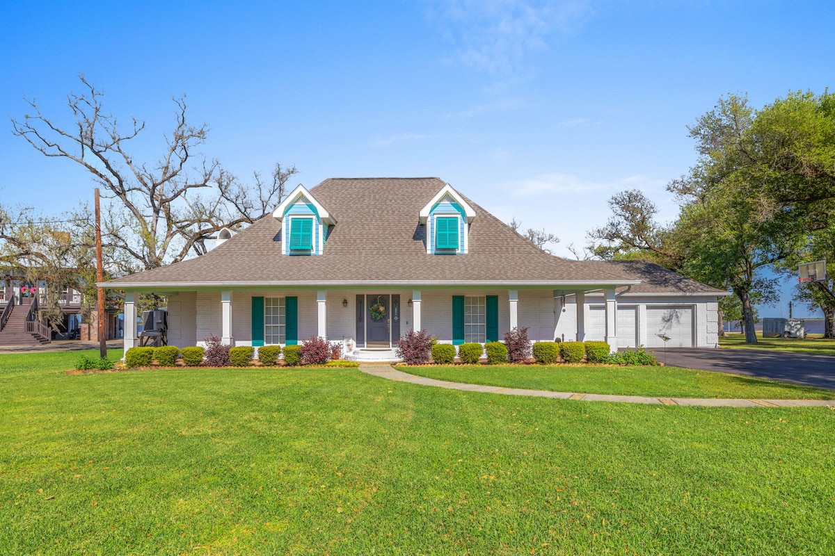 A charming two-story home features a welcoming front porch with white railings and bright turquoise shutters. Lush green lawns surround the property, which includes manicured shrubs and trees. A spacious driveway leads to the house, providing easy access to the nearby waterfront.