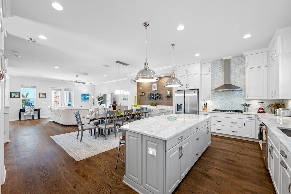 A contemporary kitchen features a large central island with a light grey finish and a white granite countertop. Sleek white cabinetry complements modern appliances. An adjacent dining area showcases a long wooden table surrounded by chairs, with an open-concept layout seamlessly connecting to the living space.