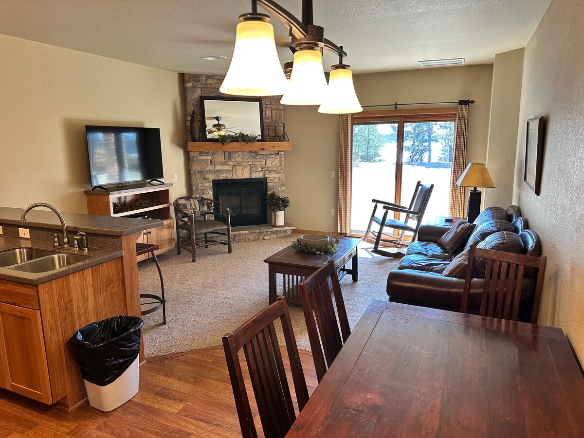 An inviting living area is featured, showcasing a gas fireplace framed by stone, a flat screen TV mounted on the wall, and a comfortable seating arrangement. A dining table is positioned nearby, with natural light streaming in from the balcony doors.