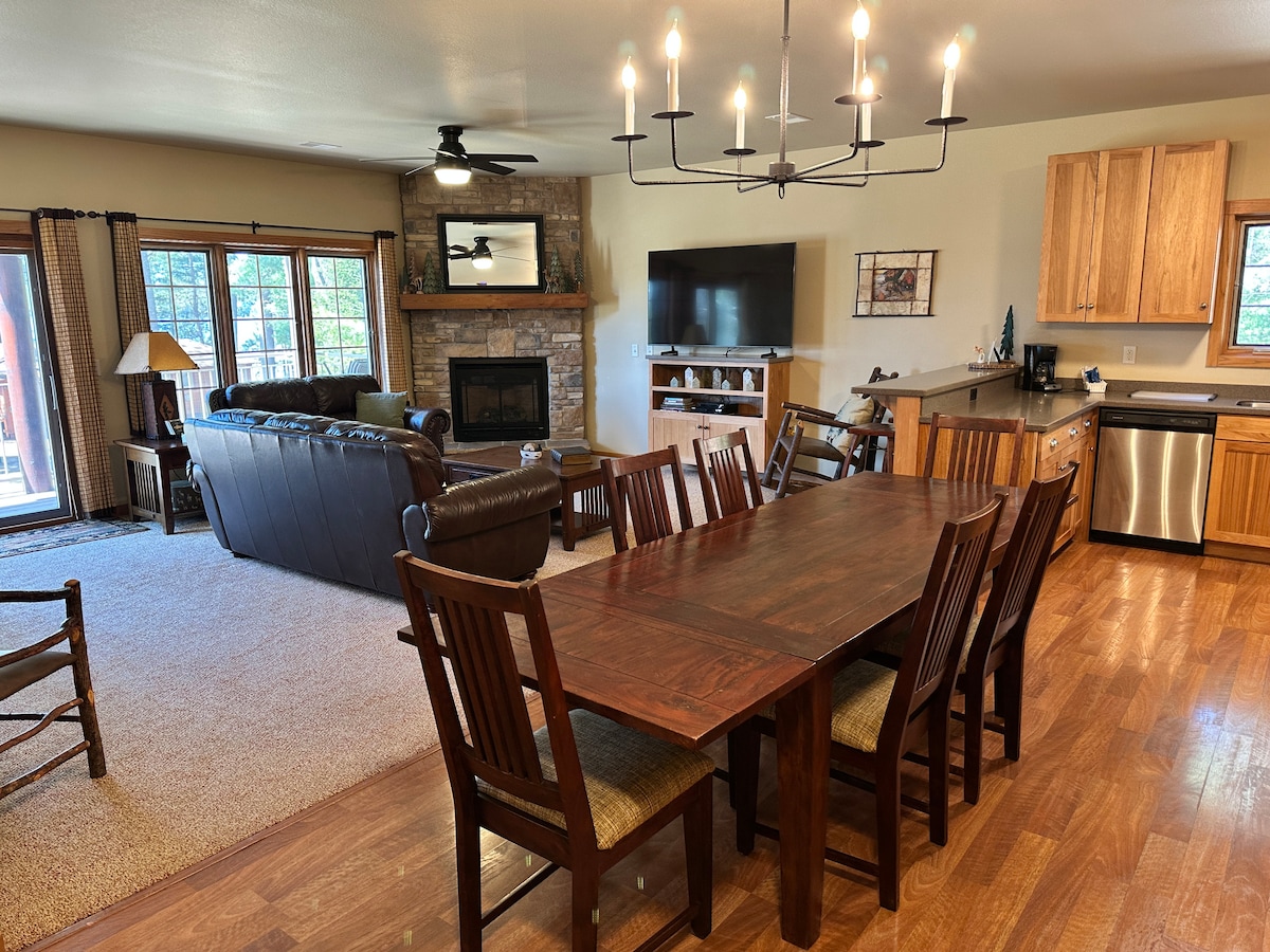 A spacious living area is visible, featuring a large wooden dining table surrounded by eight chairs. A comfortable leather sofa is arranged near a stone fireplace. Natural light fills the room through multiple windows, complemented by a flat screen TV mounted on the wall.