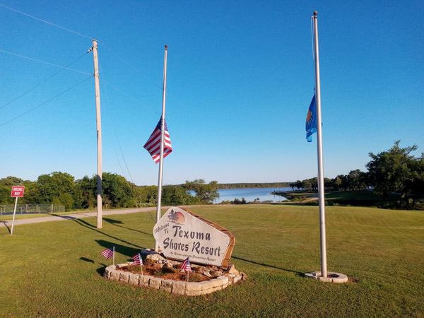 A welcoming entrance to Texoma Shores Resort is marked by a prominent stone sign. Two flags stand on either side, gently billowing in the breeze. Lush green lawns extend towards a serene lake, reflecting a clear blue sky in the background.