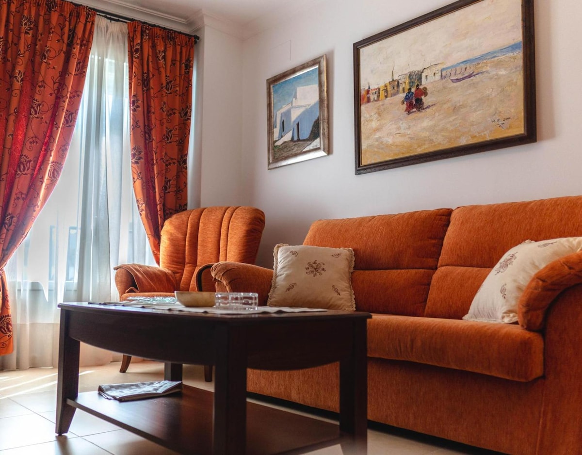 A cozy living area features comfortable orange armchairs and a matching sofa, arranged around a wooden coffee table. Natural light enters through large windows, which are framed by patterned curtains. Two framed artworks adorn the walls, adding a touch of color to the space.