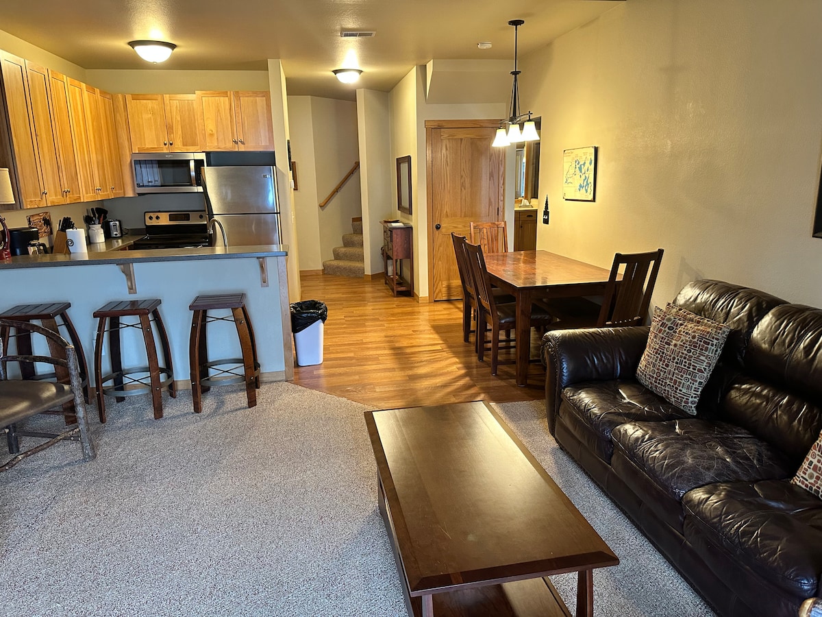 A well-lit living area features a dark leather sofa adorned with patterned cushions. A wooden coffee table sits in front, complemented by bar stools at the nearby kitchen counter. A dining area with a wooden table and chairs is visible in the background, leading to a staircase.
