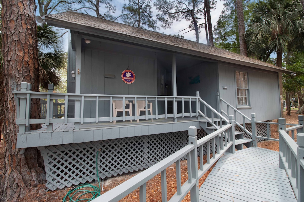 A single-story cottage features a stepping ramp leading to the entrance, providing easy access. Two simple chairs are positioned on the porch, surrounded by trees. The exterior showcases muted grey siding, complemented by white railings and lattice work underneath.
