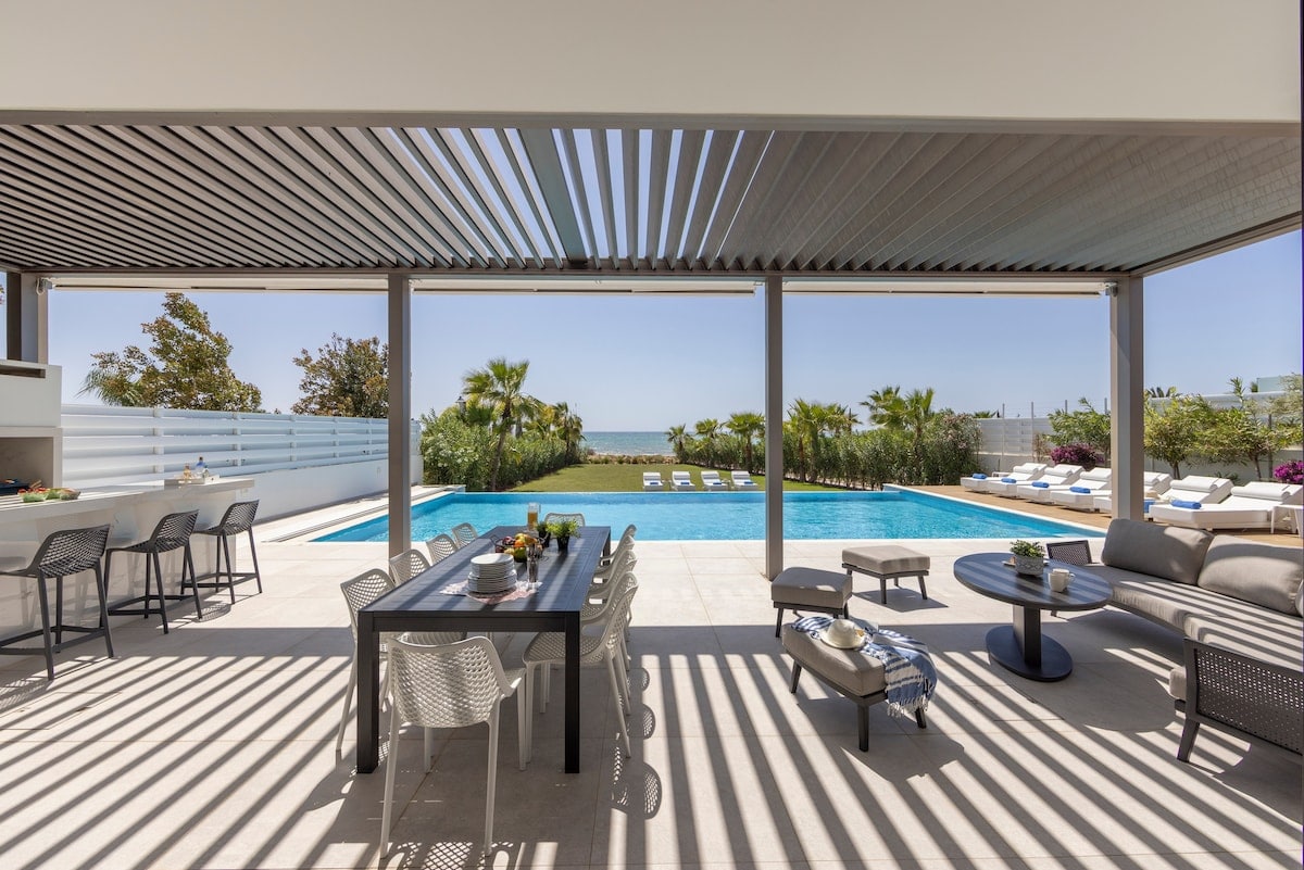 An outdoor terrace is presented, featuring a dining table accompanied by several chairs beneath a slatted roof. A spacious pool reflects the clear sky, surrounded by lounge chairs and tropical plants, creating an open and relaxed atmosphere for guests.