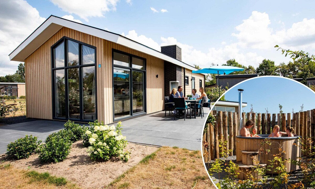 The holiday home features a modern design with large glass windows allowing ample natural light. A shaded outdoor dining area is seen, alongside a private relaxation space with a hot tub, enclosed by wooden fencing and surrounded by greenery.