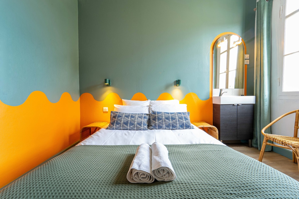 A bright bedroom showcases a double bed with white linens and decorative pillows. The walls are painted in a combination of teal and orange, creating a lively atmosphere. A small sink and mirror are positioned near the bed, while a wicker chair is placed to the side.