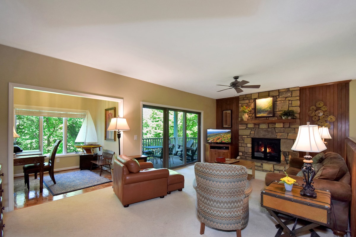 A spacious living area features a comfortable leather sofa and patterned armchair, arranged around a rustic coffee table. A stone fireplace adds warmth, while large windows invite natural light and provide views of the surrounding greenery. A dining area is visible in the background.