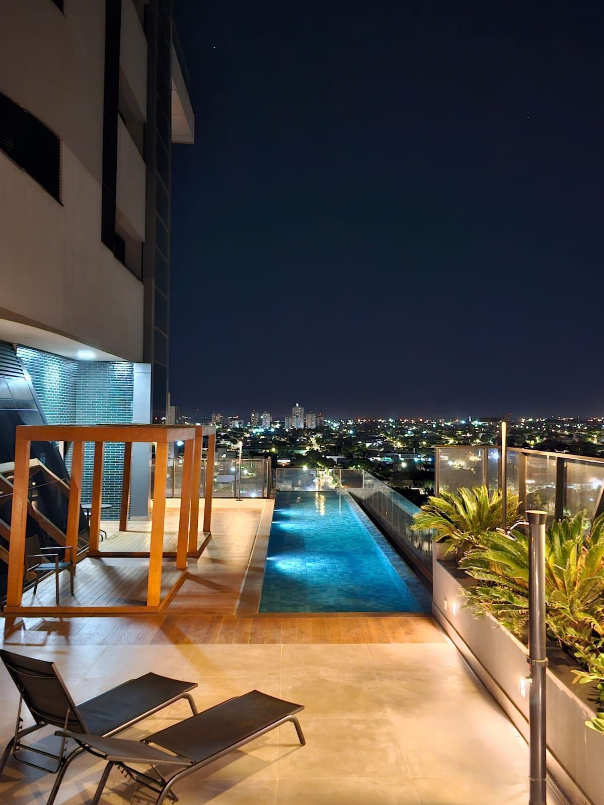 A rooftop area is shown with a tranquil swimming pool extending along the edge. Wooden lounge chairs are positioned beside the pool, which reflects the city lights at night. Lush greenery is incorporated into the space, offering a serene view of the urban skyline.