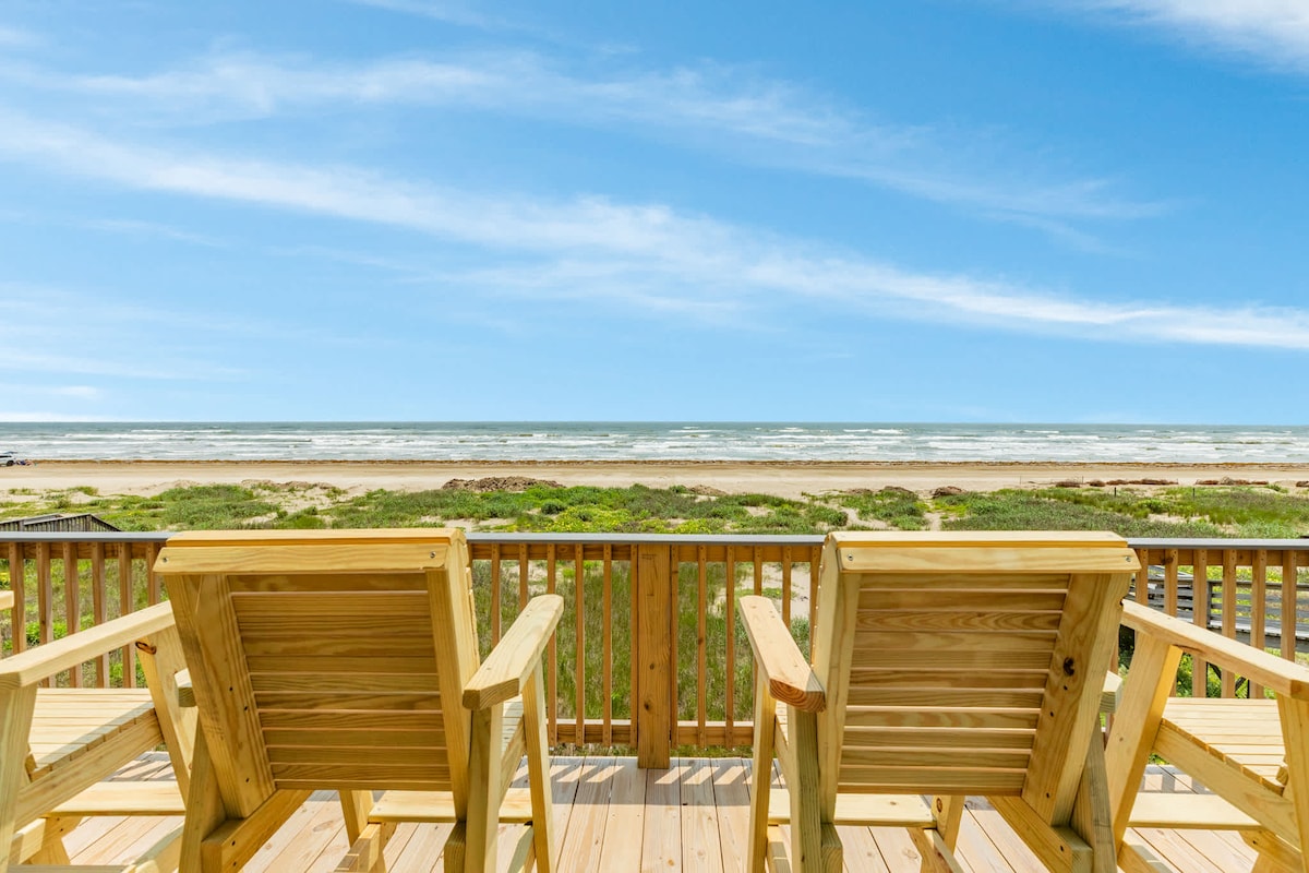 A spacious deck features two wooden chairs, positioned to overlook the beach and ocean. The expansive view reveals sandy shores and green coastal vegetation, with waves gently lapping at the shoreline under a bright blue sky.