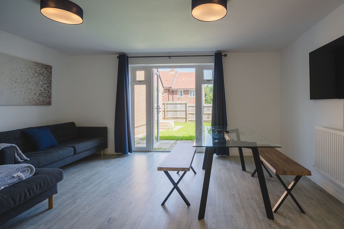 A living space is presented with a modern glass dining table and wooden benches. A comfortable grey sofa is positioned against one wall, and two light fixtures hang from the ceiling. Large glass doors lead to the shared garden, allowing natural light to fill the room.