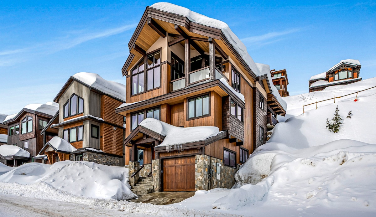 A luxurious four-storey chalet is presented, featuring a combination of wooden and stone elements. The exterior showcases large windows that offer mountain views and a covered garage. Snow blankets the surrounding area, highlighting the chalet's modern architectural design.