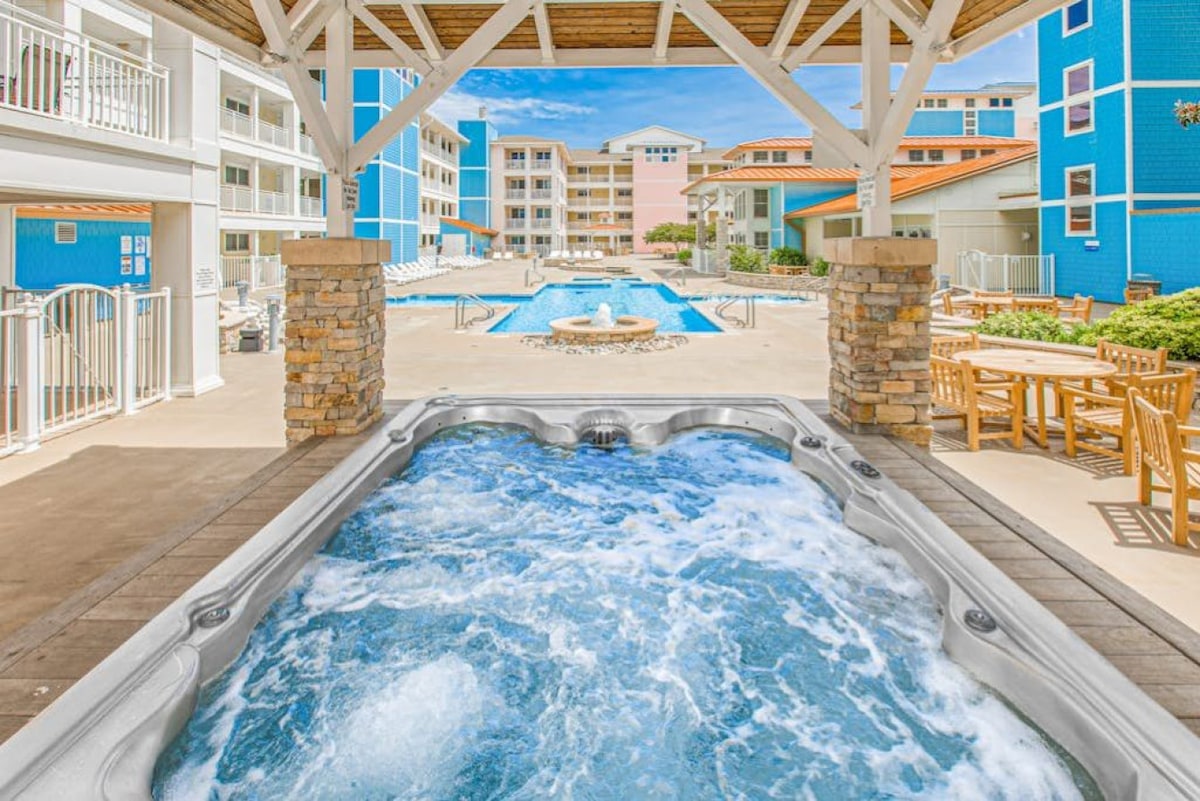 A spacious outdoor area features a hot tub with bubbling water, surrounded by a well-maintained deck. Views of multiple swimming pools and colorful buildings in the background are visible, highlighting a relaxed and inviting atmosphere for guests.