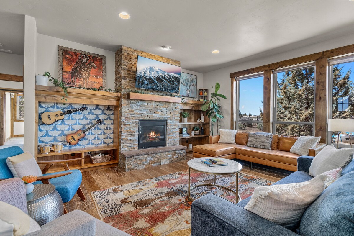 A comfortable living area is highlighted by a stone fireplace and wooden accents. Large windows allow natural light to fill the space, revealing a cozy seating arrangement with a mix of leather and fabric upholstery. Decorative artwork and a patterned area rug add warmth to the environment.