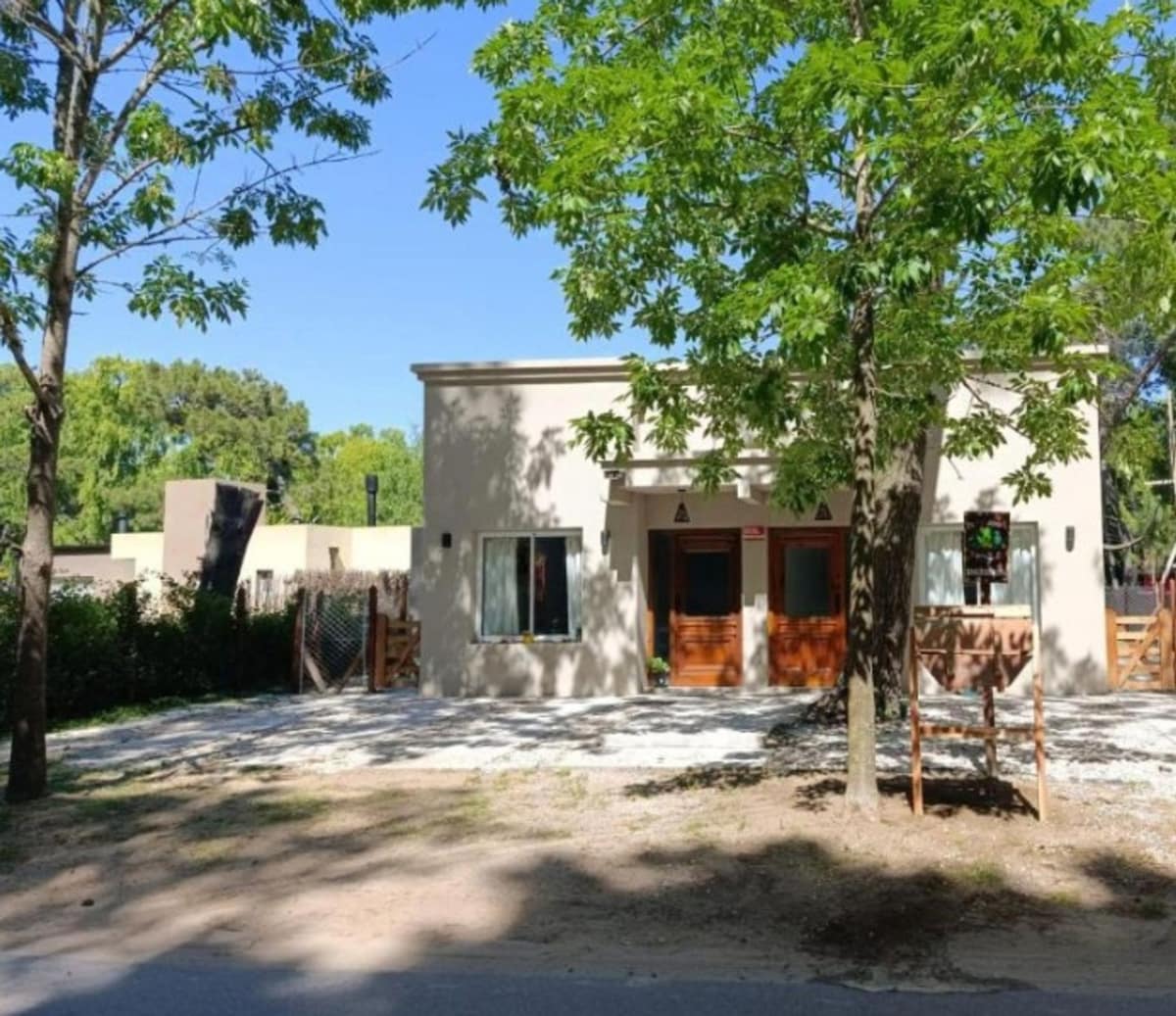 A modest single-story house is shown, framed by lush green trees. The entrance features two wooden doors flanked by large windows. A gravel path leads to the house, surrounded by a natural landscape that enhances the inviting setting.