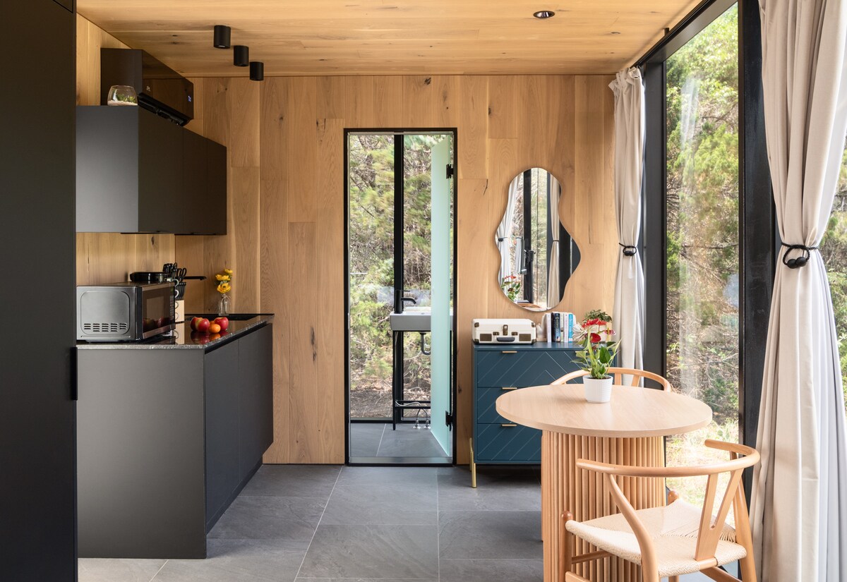 A modern kitchen area features sleek black cabinetry and stainless-steel appliances, complemented by light wood paneling on the walls. A round dining table with natural wood chairs sits invitingly, while large windows bring in natural light and offer views of the surrounding greenery.