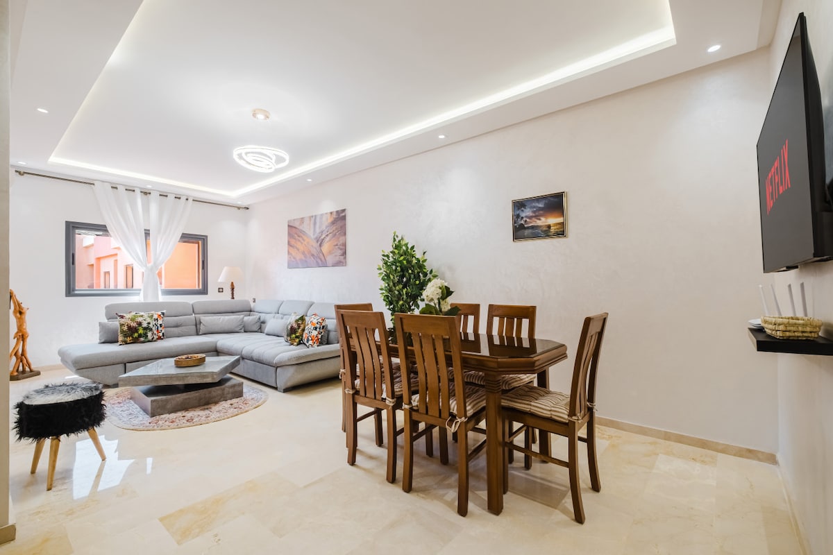 The spacious living area is furnished with a large sectional sofa and a wooden dining table surrounded by six chairs. Natural light enters through the window, which is complemented by decorative wall art. The soft hues and modern decor create a comfortable space for relaxation.