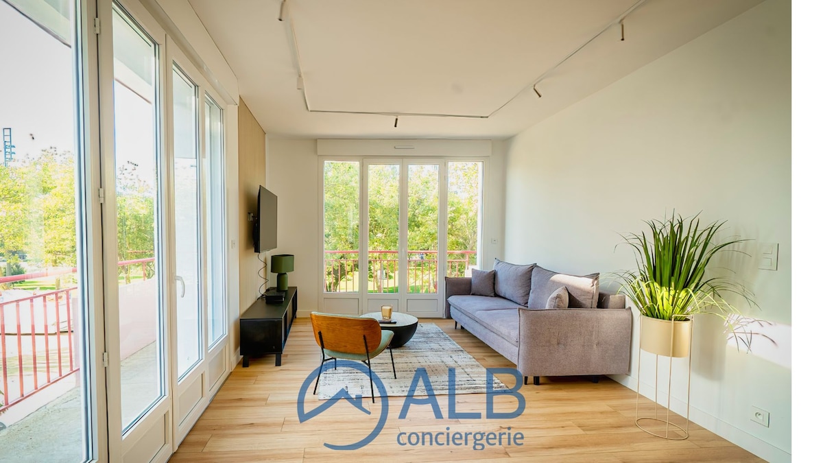 A bright and airy living space features large glass doors that provide natural light and a view of lush greenery. A comfortable sofa sits adjacent to a minimalistic coffee table, while a flat-screen TV is mounted on the wall opposite. A leafy plant adds a touch of nature to the room.