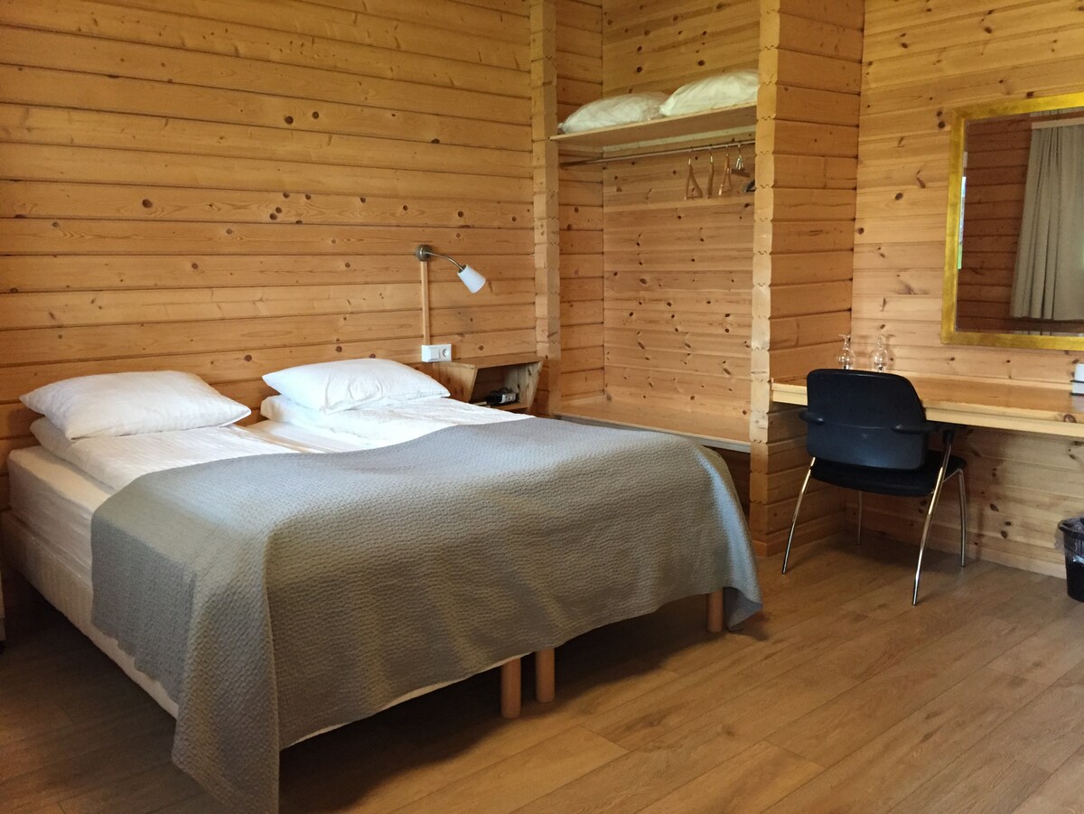 A wooden interior defines the sleeping area, featuring a spacious bed dressed in neutral bedding. Above the bed, storage shelves hold additional pillows. A small desk with a chair is positioned nearby, illuminated by a wall-mounted lamp, enhancing the cozy ambiance.