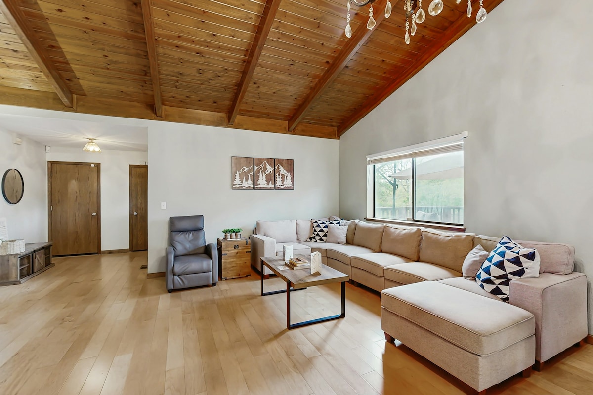 A spacious living room features a high wooden ceiling and a large sectional couch adorned with decorative pillows. Natural light enters through a window, illuminating a coffee table in the center. A recliner is positioned nearby, and two doors lead to other areas of the home.