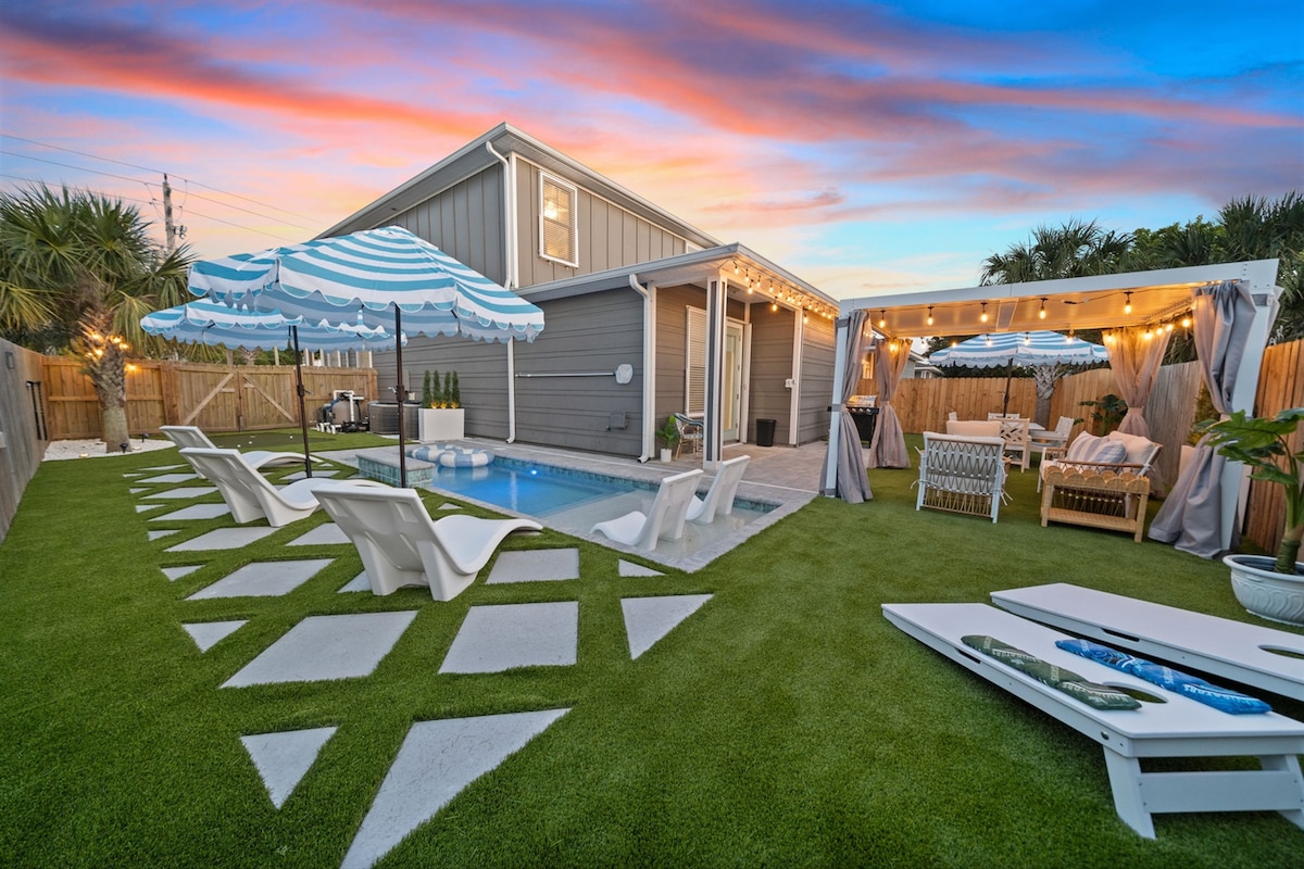 An outdoor space features a private inground pool, surrounded by lounge chairs and umbrellas. A deck with a covered pergola offers seating and dining options, illuminated by string lights. Lush green grass and palm trees create a tranquil atmosphere with a colorful sunset in the background.