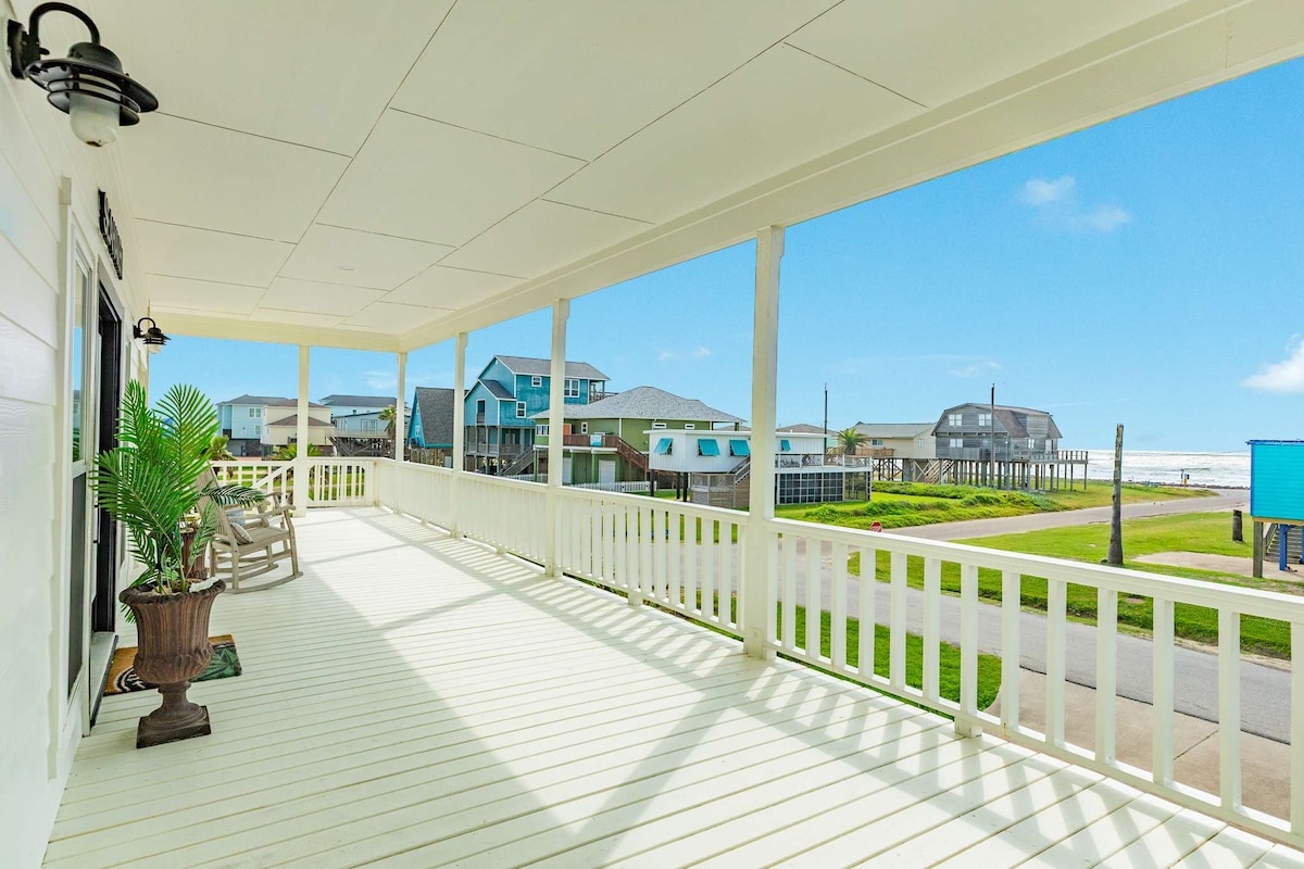 An expansive wrap-around deck features white wooden flooring and sturdy railings, providing clear ocean views. Comfortable seating arrangements invite relaxation, with well-maintained greenery adding a touch of nature. Sunlight brightens the scene, creating a welcoming outdoor space for enjoying coastal breezes.
