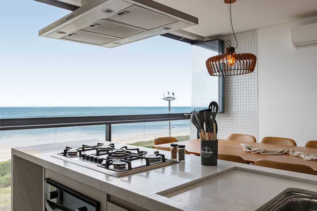 A modern kitchen is highlighted, featuring a sleek gas stove and an array of kitchen utensils in a caddy. The large window offers a panoramic view of the ocean, enhancing the dining area visible in the background with wooden chairs around a table.