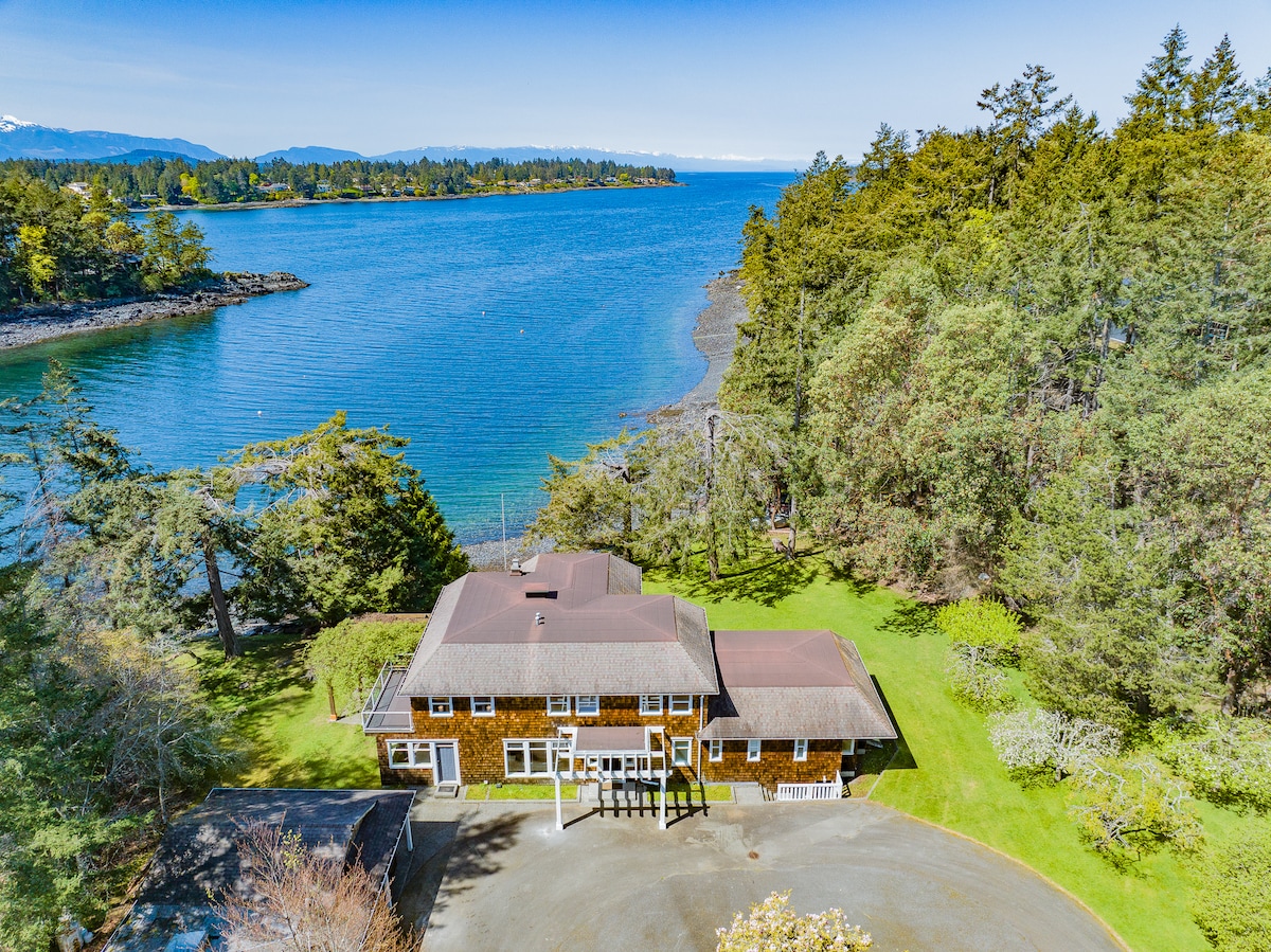 An aerial view shows a two-story cedar-shake home nestled in lush greenery along the shores of Nanoose Bay. The expansive lawn leads to rocky shoreline, with calm blue waters reflecting the sky and surrounding forested hills in the background.
