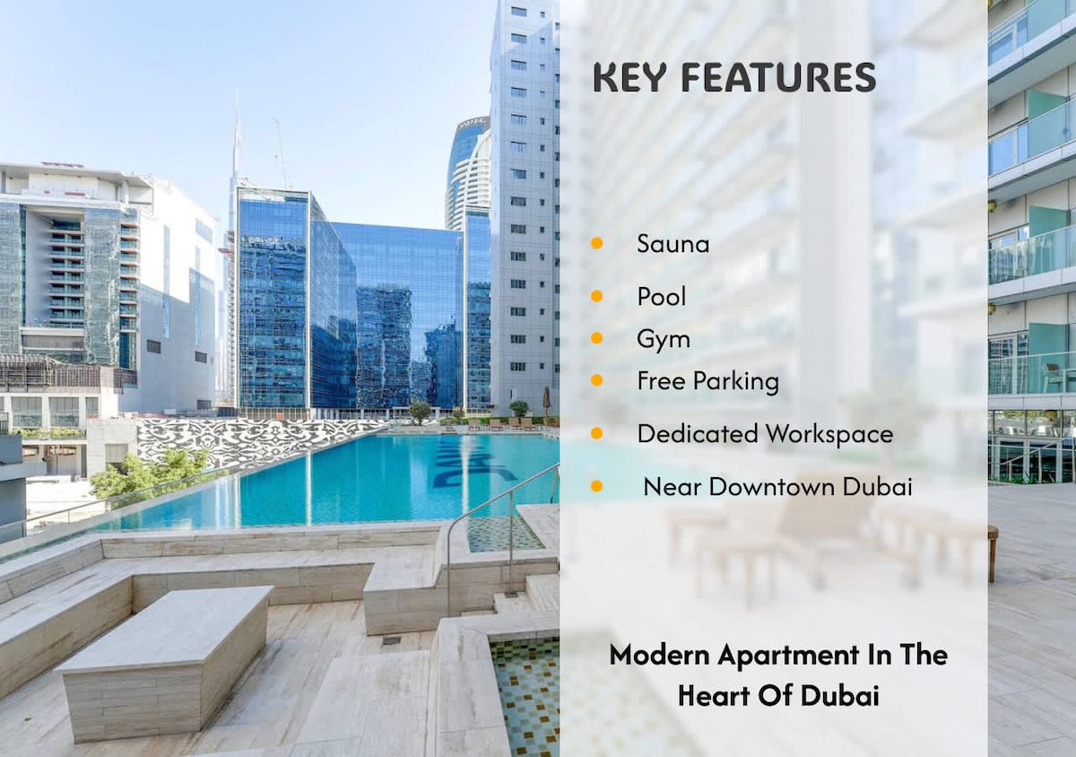 The image features a large outdoor swimming pool surrounded by stylish lounge areas and glass-fronted buildings. A modern fitness center is visible nearby, along with a well-maintained landscaping. Sunlight reflects off the water, providing a serene view in a vibrant urban space.