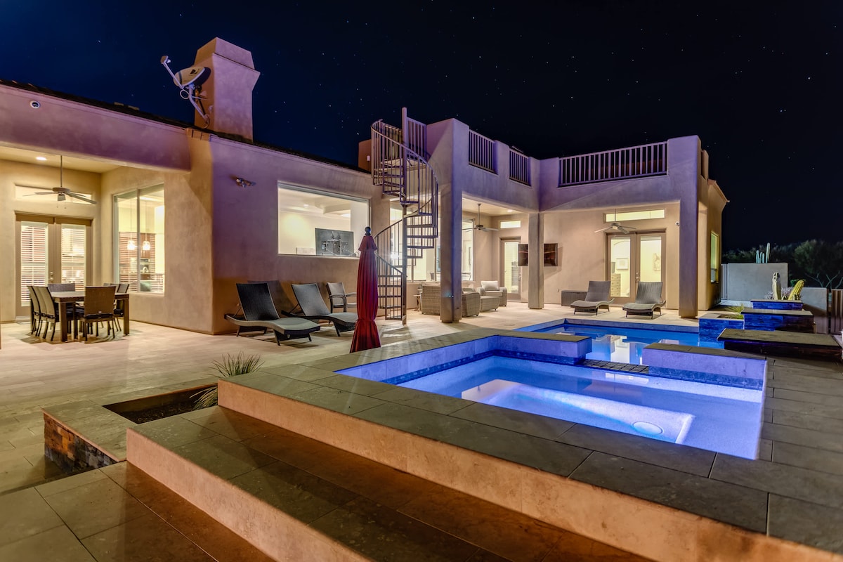 The outdoor area is highlighted by a heated pool and hot tub, illuminated with calming blue lights. A rooftop terrace is accessible via a spiral staircase, and a dining table is situated nearby under the evening sky, creating a serene atmosphere for relaxation.