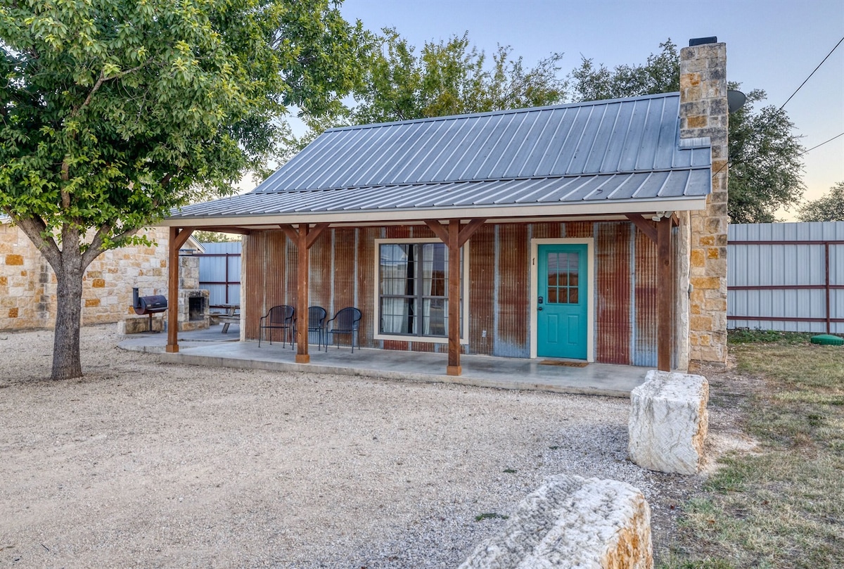 A charming cabin exterior is visible with a metal roof and a turquoise front door. The porch is furnished with four chairs, and the surrounding area is landscaped with gravel and a large tree. A fire pit is positioned in the side yard.