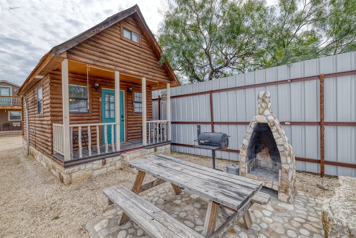 A charming log cabin exterior is highlighted, featuring a front porch with seating and a stone fire pit area. A picnic table is positioned on a gravel surface, with a charcoal BBQ pit nearby. Surrounding the cabin is a secure metal fence, providing privacy.