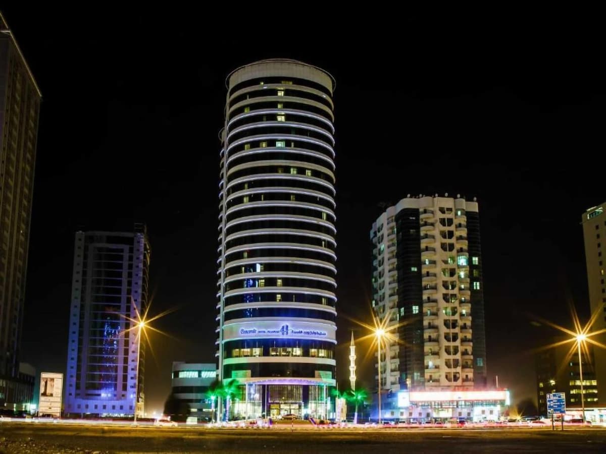 The building stands tall at night, featuring a modern circular design with illuminated lines and features. Surrounding towers reflect the vibrant city lights, contributing to the lively urban atmosphere. Streetlights illuminate the foreground, enhancing visibility along the busy road.