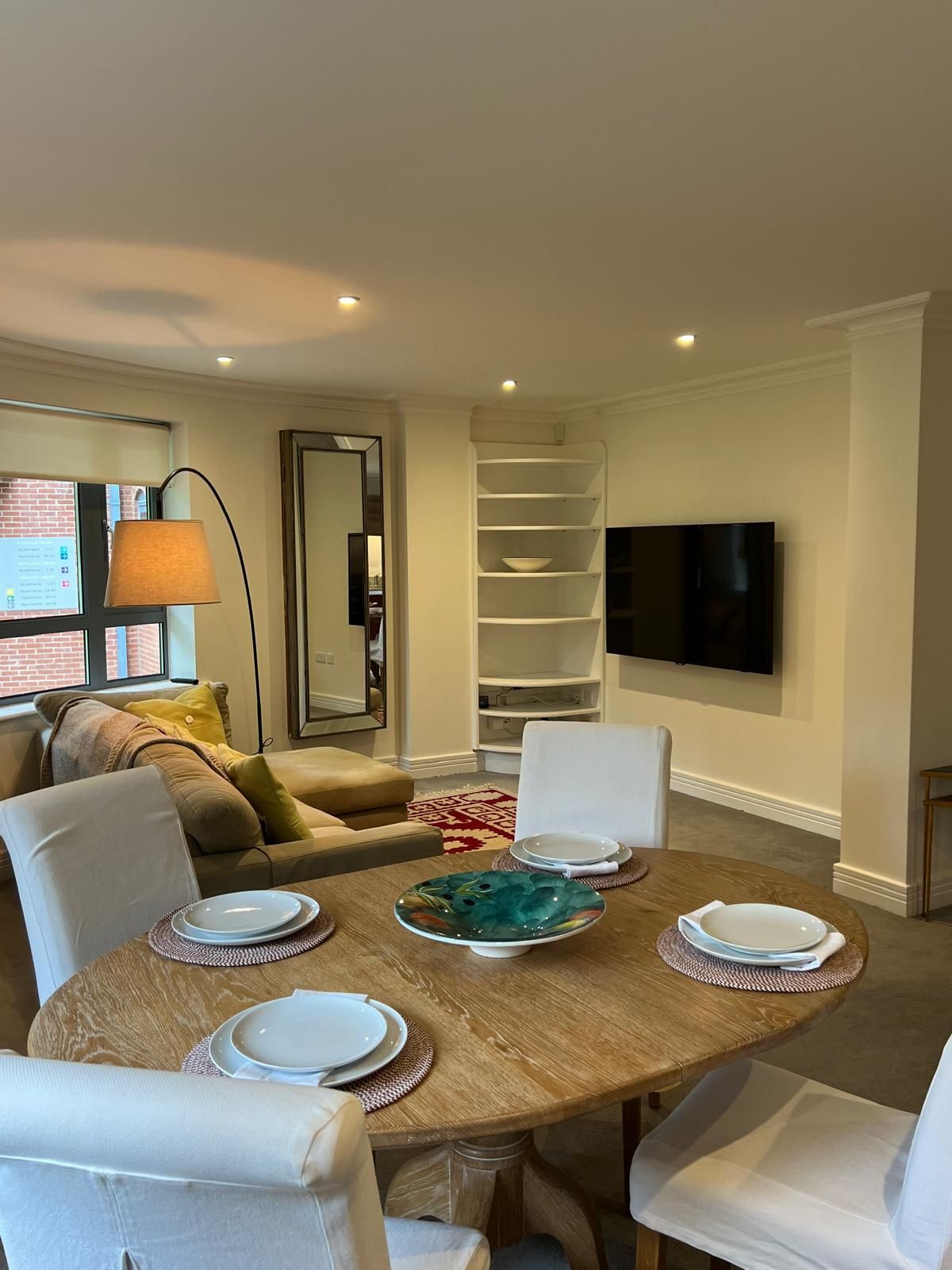 An inviting open-plan living and dining area features a round wooden table set for four, with white plates and a colorful dish at the center. A comfortable sofa, accented by green and beige cushions, is positioned next to a large wall-mounted television. Soft lighting enhances the space.