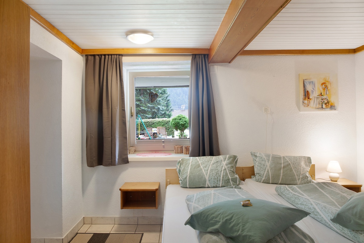 A serene bedroom is presented with two beds arranged side by side, adorned with light green bedding. Natural light enters through a window with curtains, illuminating the room. A small bedside table holds a lamp, while a decorative piece is hung on the wall.