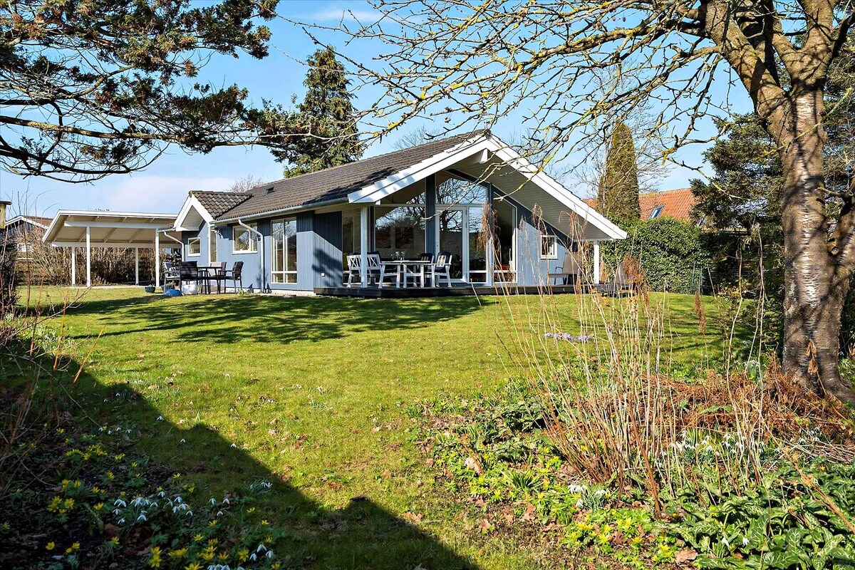 The modern cottage is surrounded by a lush garden with vibrant greenery and blooming flowers. Large windows reflect natural light, and the outdoor terrace features seating for relaxation. The property is framed by tall trees, creating a serene atmosphere near the beach.