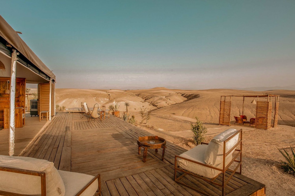A spacious wooden terrace offers comfortable seating with neutral cushions, overlooking the vast Agafay Desert. The undulating sands stretch into the horizon, accented by distant rocky hills. A small outdoor dining area is visible in the background, creating a peaceful setting for relaxation.